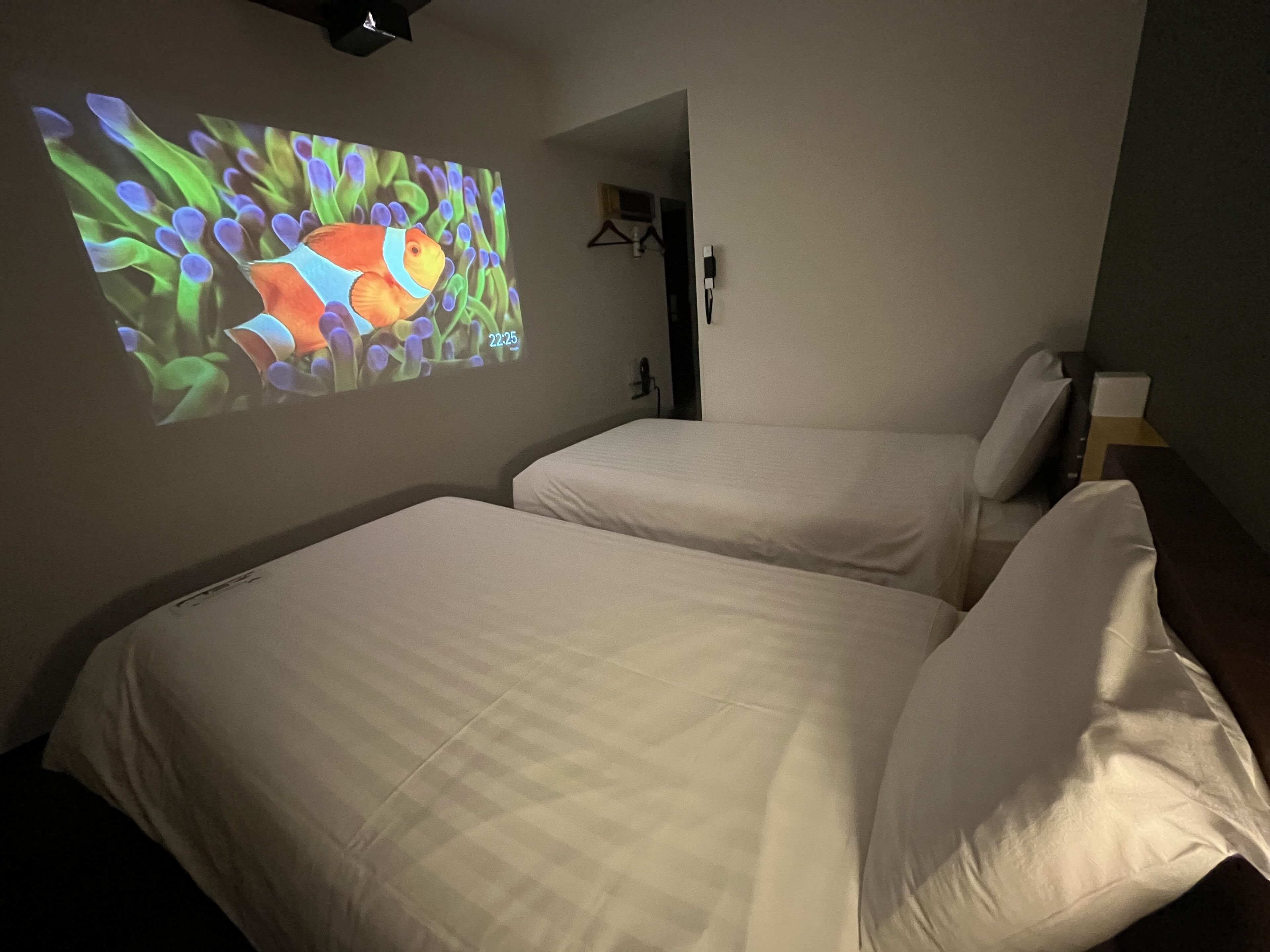 A hotel room with two beds, a wall-mounted projector displaying a colorful image, and a door leading to another room.