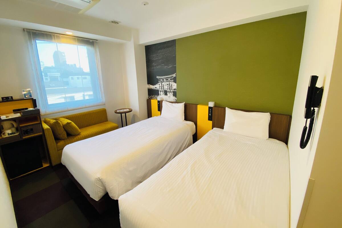 Ein Hotelzimmer mit zwei Betten, einem Schlafsofa, einem kleinen Tisch, einem Telefon und einem Fenster mit Blick auf Gebäude.