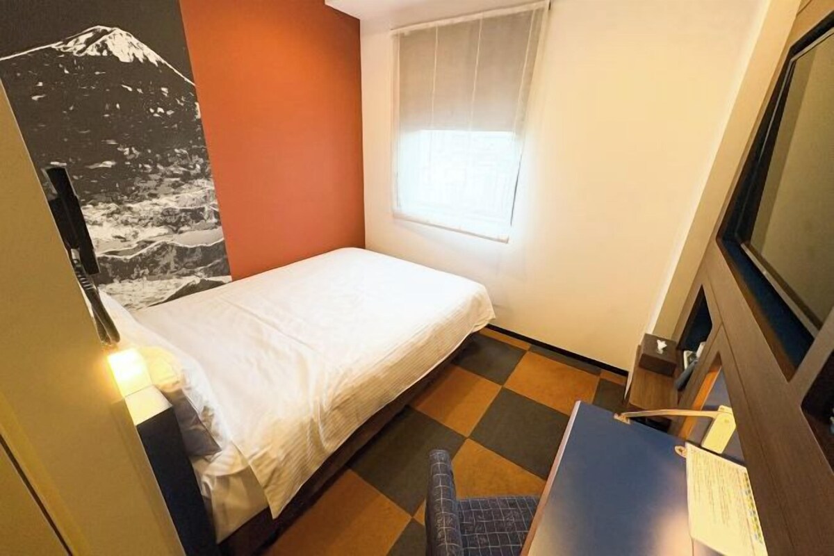 A small, single-bed room with a checkered floor, a window with blinds, and a wall-mounted TV.
