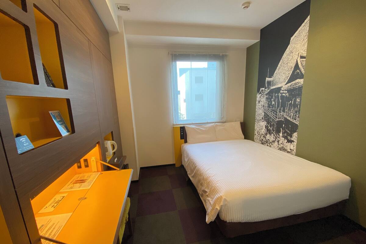 Ein Hotelzimmer mit einem Bett, einem Schreibtisch mit Stuhl, einem Fenster und einer Wand mit Stadtbild-Mural.