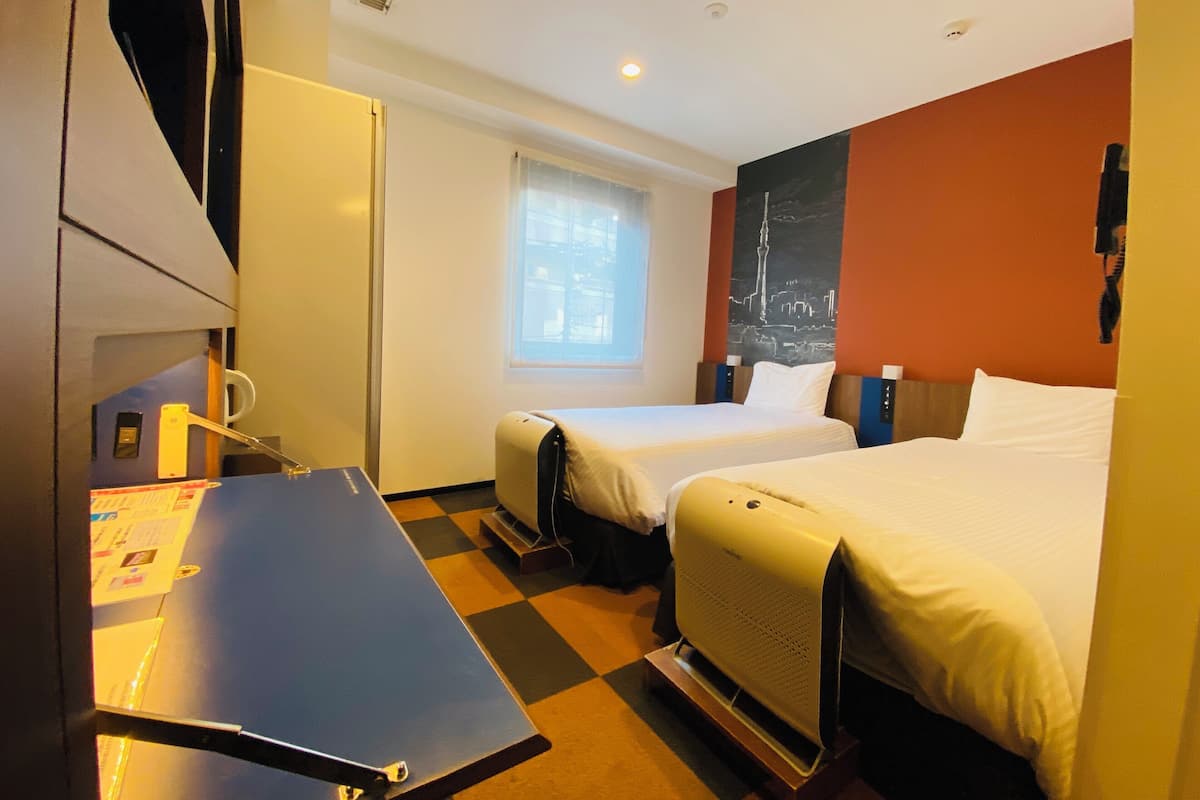 Ein Hotelzimmer mit zwei Betten, einem Schreibtisch, einem Stuhl, einem Fenster und einer Wand mit Stadtbild-Kunstwerk.