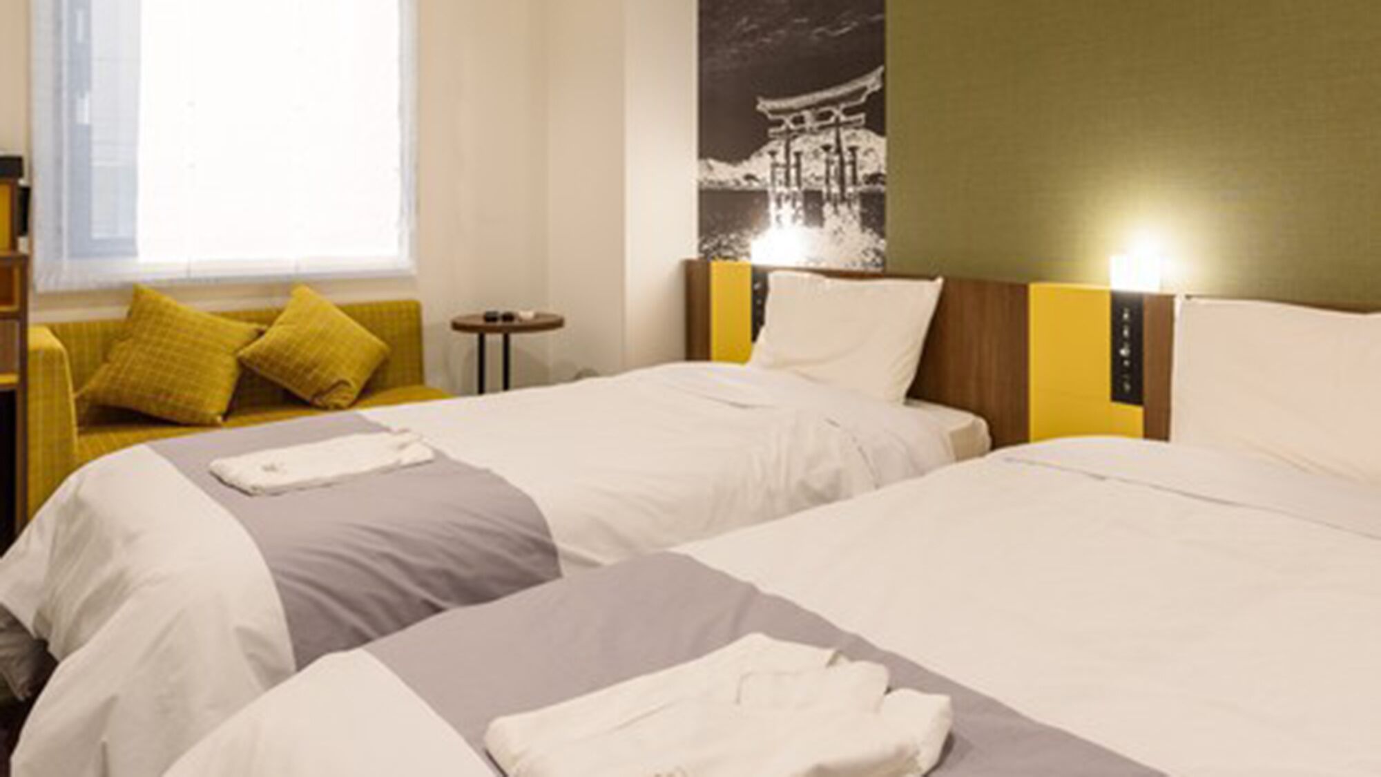 A hotel room with two beds, a sofa, a small table, and a wall-mounted light.