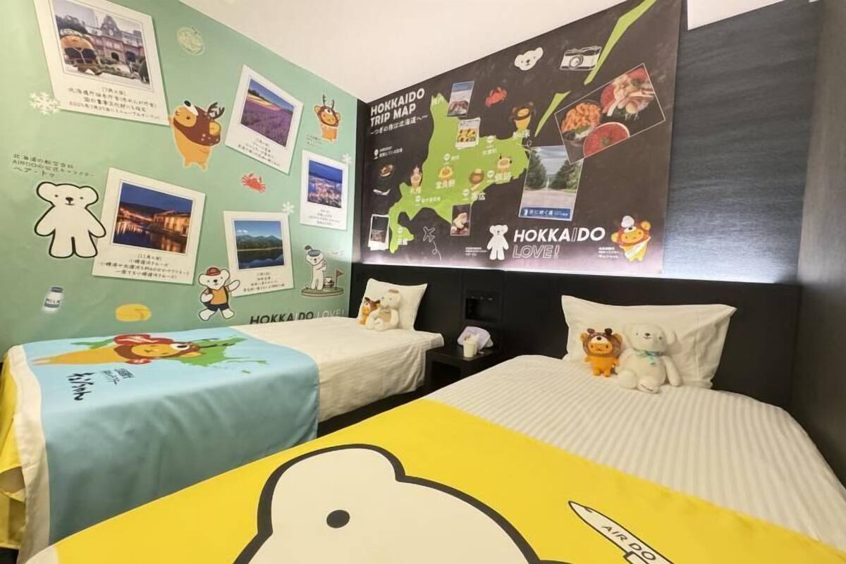 Ein Hotelzimmer mit zwei Betten, jedes mit einer andersfarbigen Bettdecke, einer mit Reisefotos und Aufklebern verzierten Wand und einer großen Karte an der Wand.