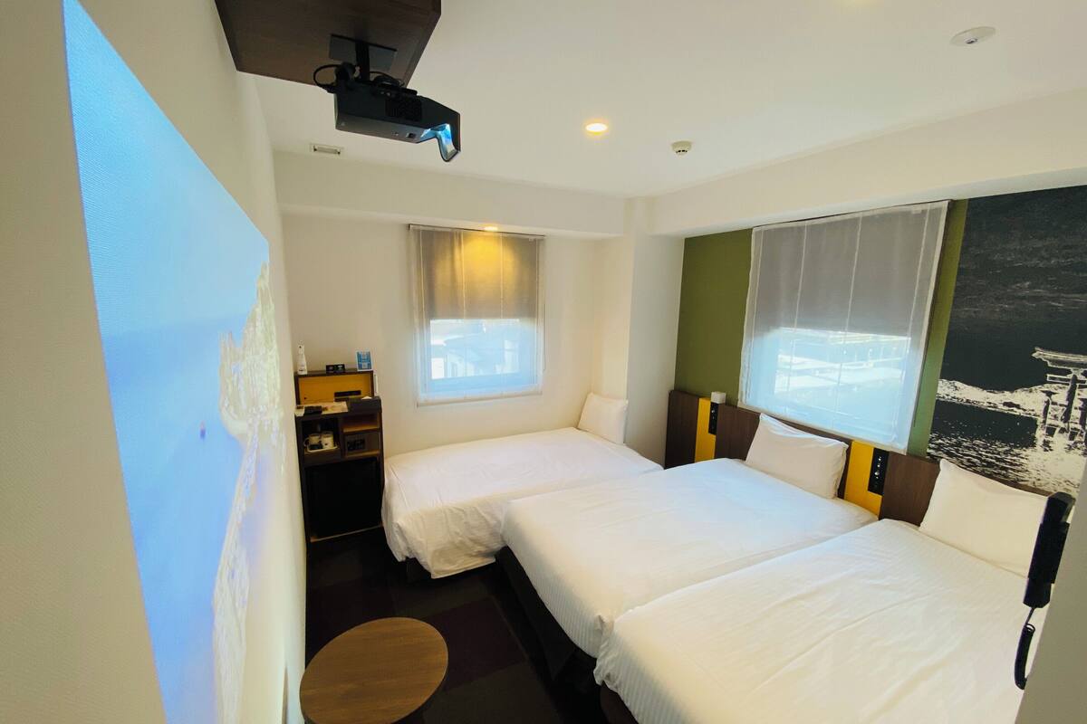 Ein Hotelzimmer mit zwei Betten, einem kleinen Tisch, einem Schreibtisch mit Stuhl, einem Projektor und einem Fenster mit Blick.