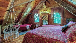 Un dormitorio acogedor en el ático, con techo inclinado, una silla colgante, cama con colcha estampada de rojo y cabecera de madera.