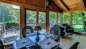 Una terraza de madera con mesa y sillas, un ventilador de techo y vista al bosque.