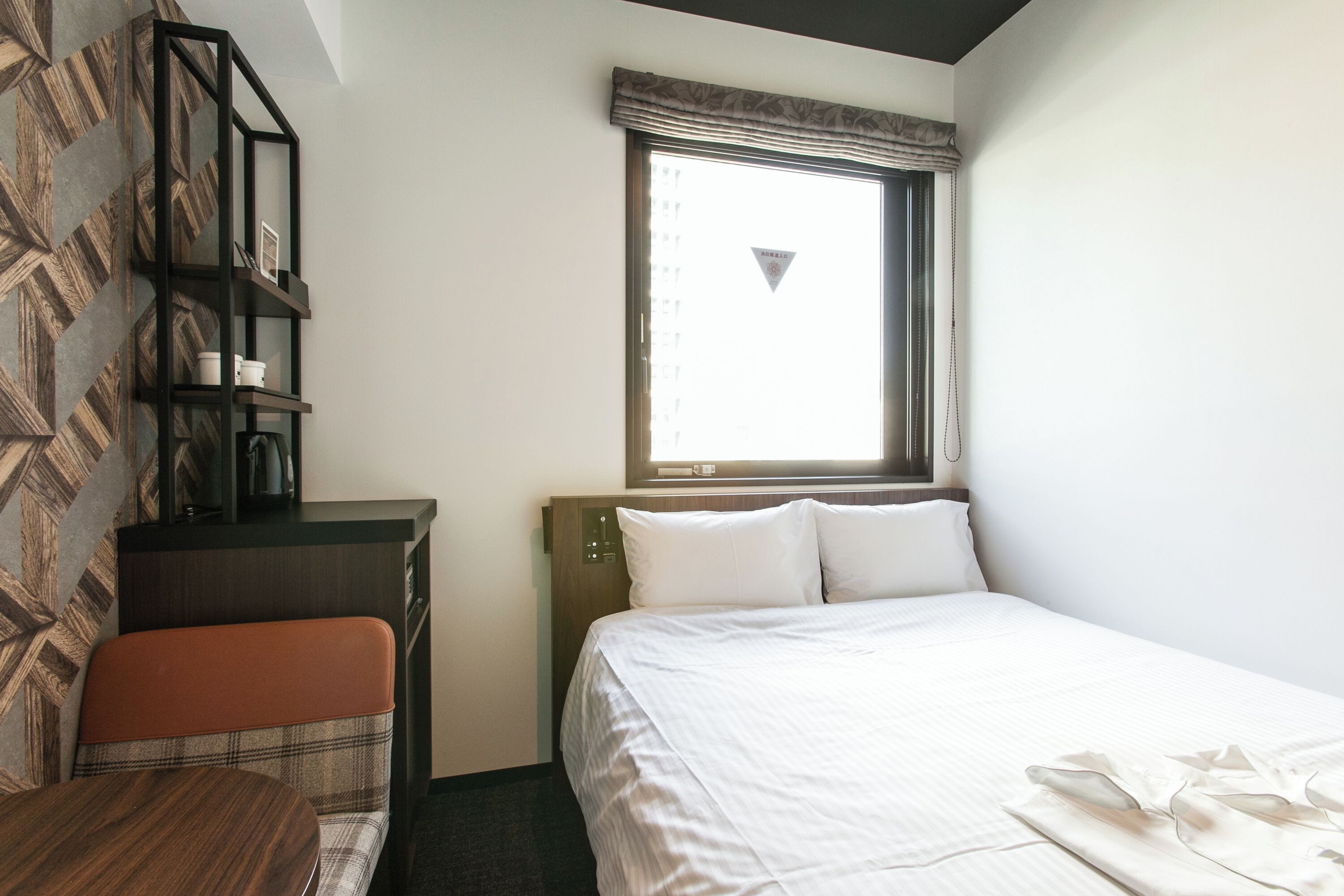 A hotel room with a bed, a desk with a chair, a bookshelf, and a window with a view.