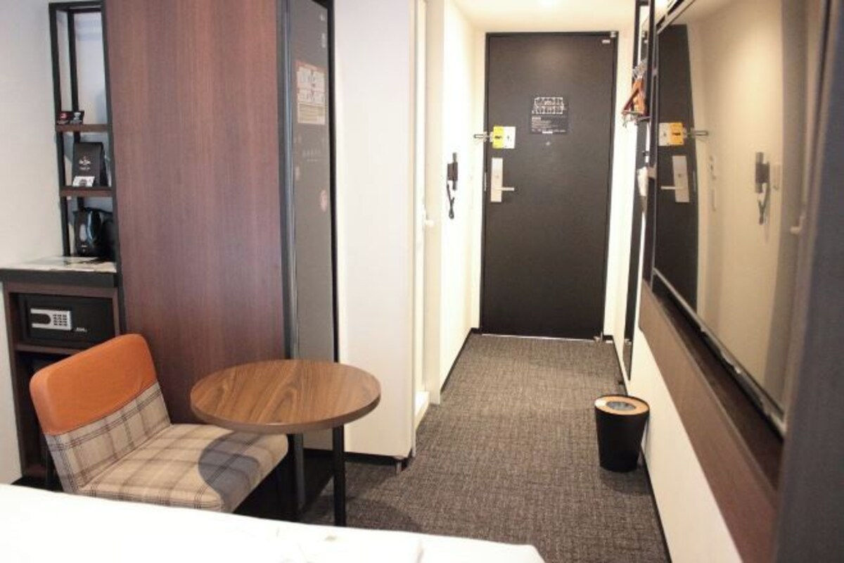 Quarto de hotel com cama, mesinha, cadeira, guarda-roupa e porta.