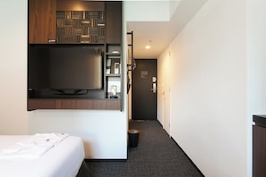 A hotel room with a bed, a TV mounted on the wall, a small desk with a phone and a lamp, and a trash bin.