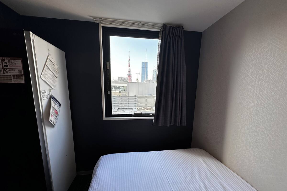 Una habitación de hotel con cama, ventana con cortina, puerta y pared con un tablón de anuncios.