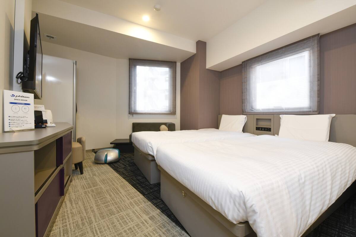 Una habitación de hotel con cama, escritorio, silla, mesita, cubo de basura y ventana.
