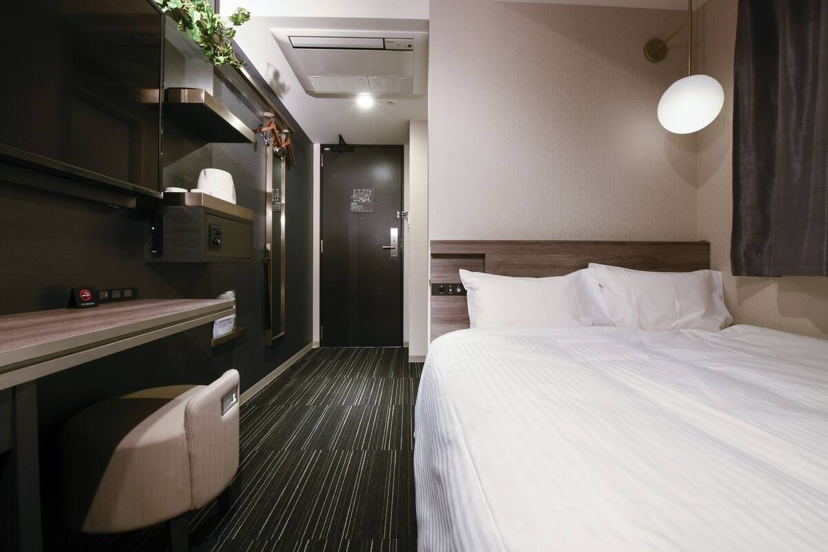 Una habitación de hotel con cama, escritorio, silla, una kitchenette pequeña y una puerta.
