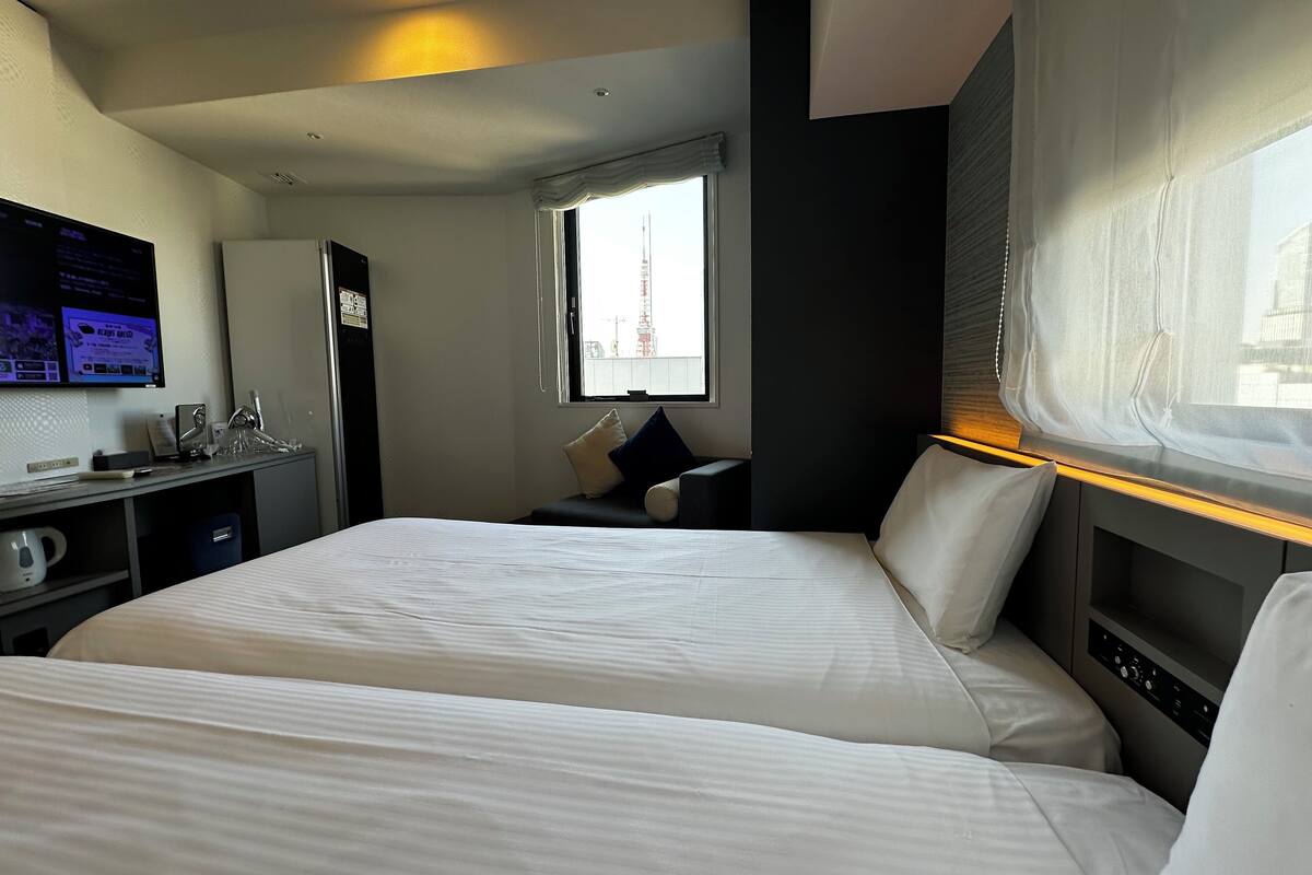 Habitación de hotel con dos camas, un escritorio con computadora y tetera, una ventana que ofrece vista a una torre, y un panel de control en el cabecero de la cama.
