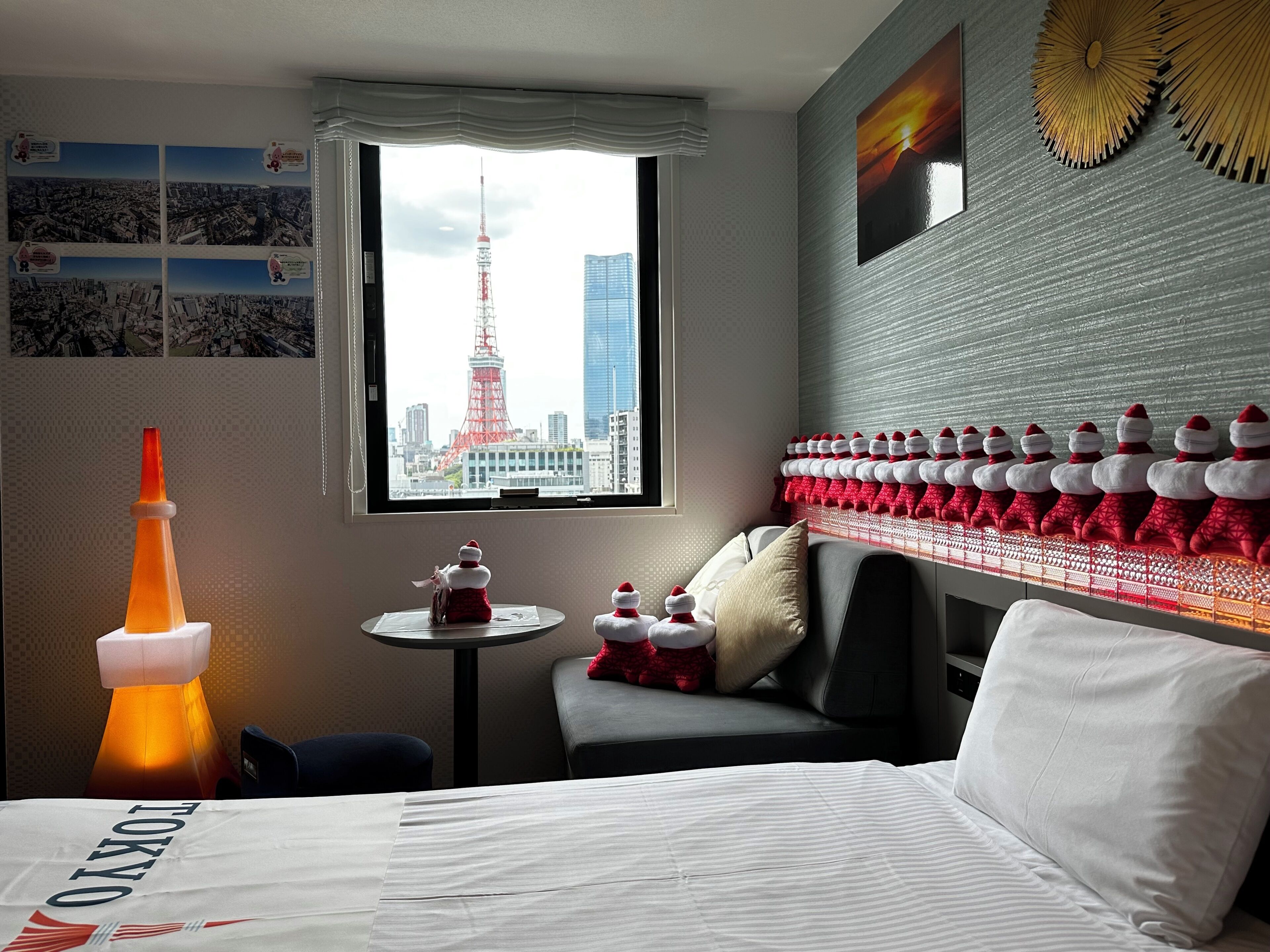 A hotel room with a bed, a small table, a window with a cityscape view, and a wall decorated with red and white pillows.