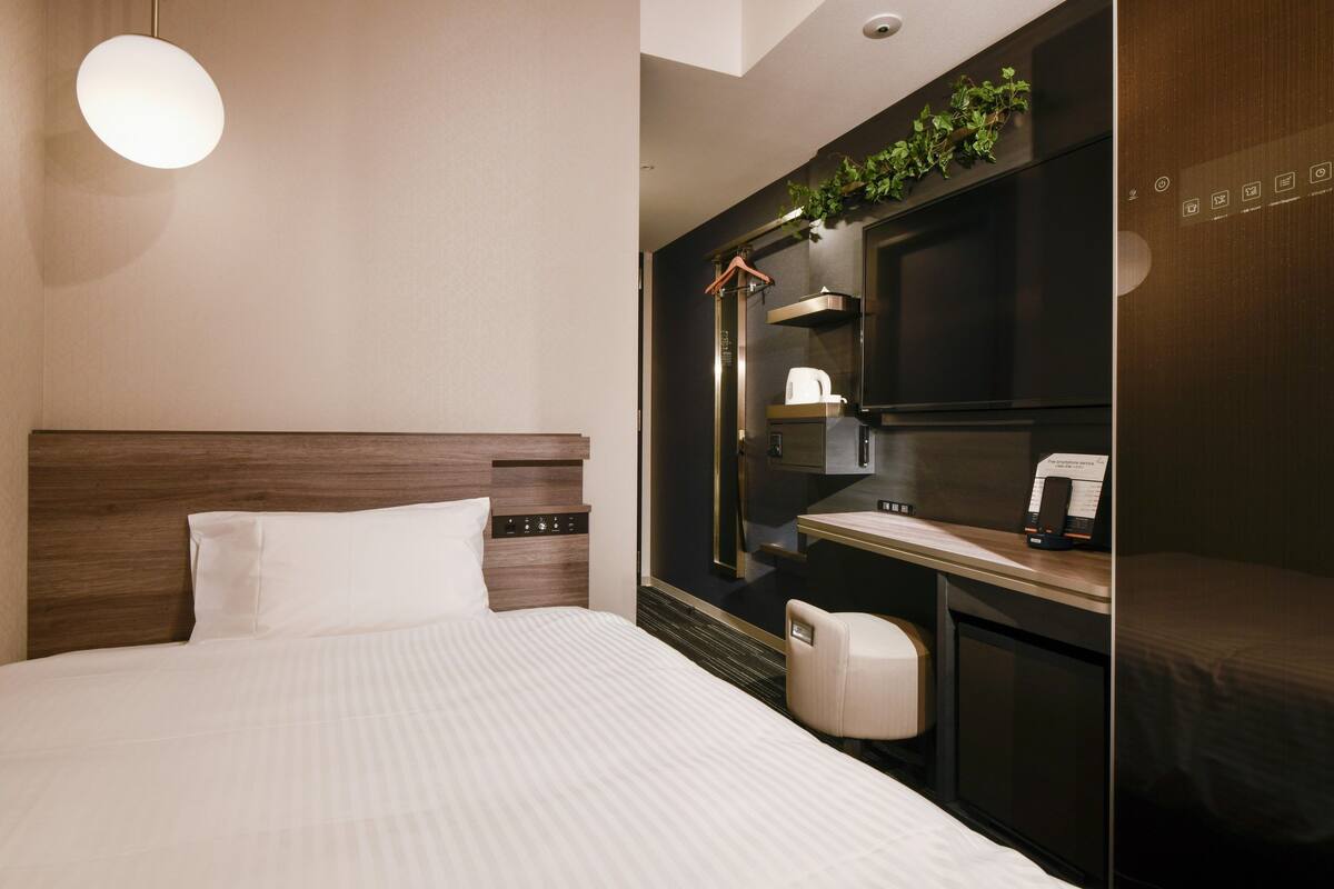 Una habitación de hotel moderna con una cama grande, un escritorio con silla, televisor y una pequeña kitchenette.