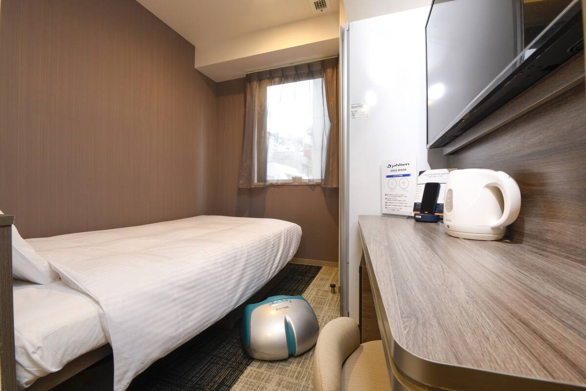 Habitación de hotel con una cama individual, un escritorio pequeño con tetera, cafetera y una silla de masajes.