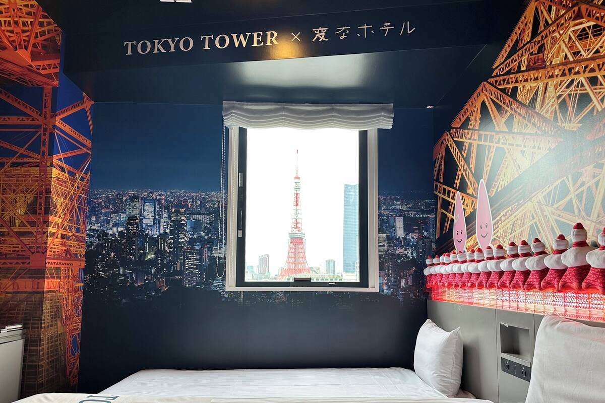 Habitación de hotel con un ventanal grande que ofrece vistas a la ciudad, incluyendo una torre destacada. La habitación tiene una cama con cubrecama y almohadas blancas, una mesita de noche y una pared con un diseño decorativo.