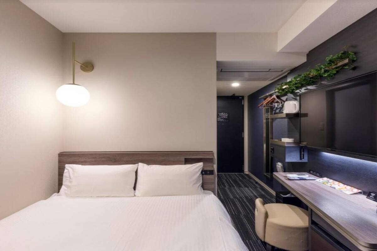 Habitación de hotel con cama, escritorio, silla, estante empotrado con plantas y una puerta.
