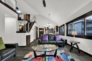 A spacious living room with a glass coffee table, a leather sofa, and a colorful rug. A staircase leads to an upper level, and there is a large window providing a view outside.