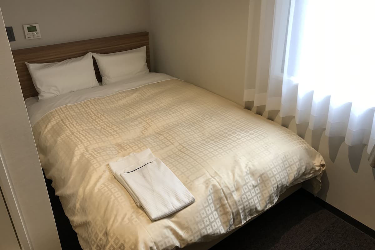 Een netjes opgemaakt bed met een beddekleed met patroon, een opgevouwen witte handdoek en een thermostaat aan de muur.