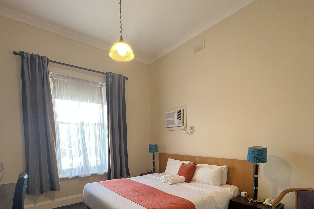 Hotellihuone, jossa on sänky, työpöytä, tuoli, lamppu ja ikkuna, jossa on verhot.