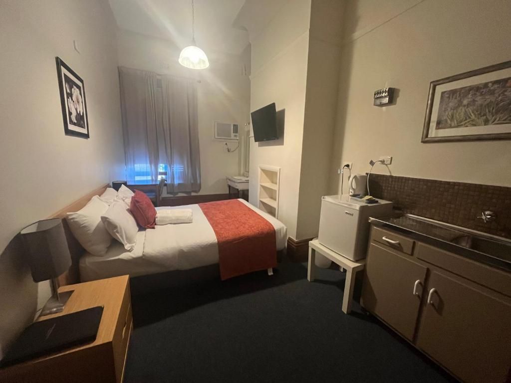 A hotel room with a bed, bedside table, a desk with a chair, a kitchenette, a TV, and a window with curtains.