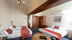 Habitación de hotel con dos camas, una mesa redonda de madera, una silla, un escritorio con lámpara, un armario y un cuadro en la pared.