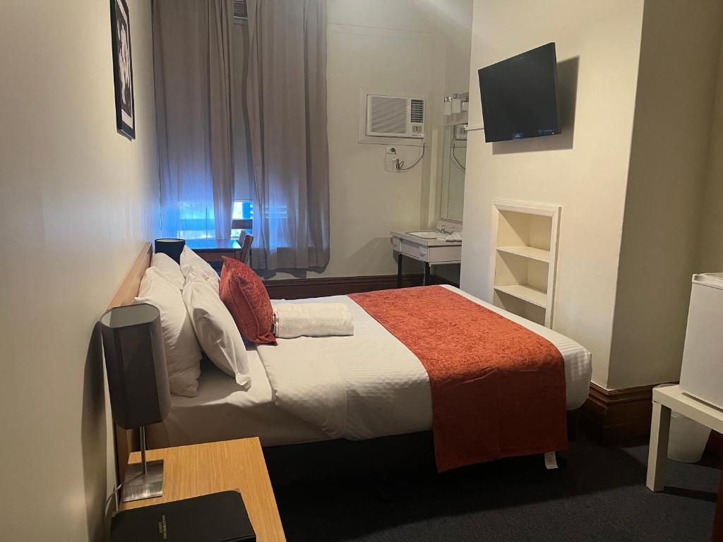 A hotel room with a bed, a bedside table, a TV mounted on the wall, a window with curtains, and a bathroom with a sink and mirror.
