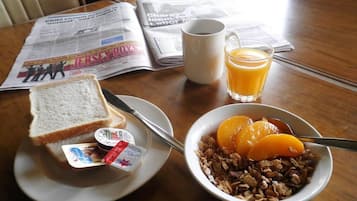 Desayuno con tostadas, cereal, jugo de naranja y un periódico sobre una mesa de madera.
