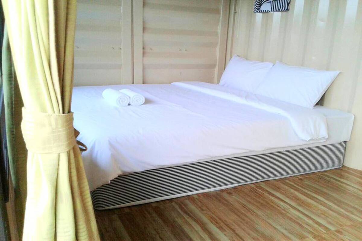 A bed with white linens and a rolled towel, a wooden floor, and a curtain partially visible.