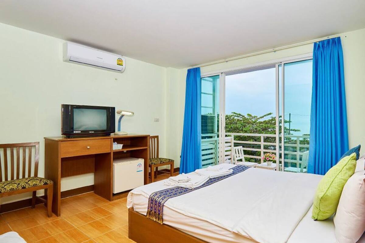 A hotel room with a bed, a desk with a television, a chair, and a balcony with blue curtains.