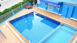 A swimming pool with a wooden deck, a small seating area, and a blue building with a tiled roof. - Blue House Beach (Pattaya)