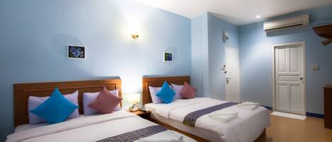 Habitación de hotel con dos camas, cada una con cabecero de madera, ropa de cama blanca y almohadas decorativas. Se observa una mesita de noche con una lámpara, un aire acondicionado de pared y una puerta con pomo blanco.