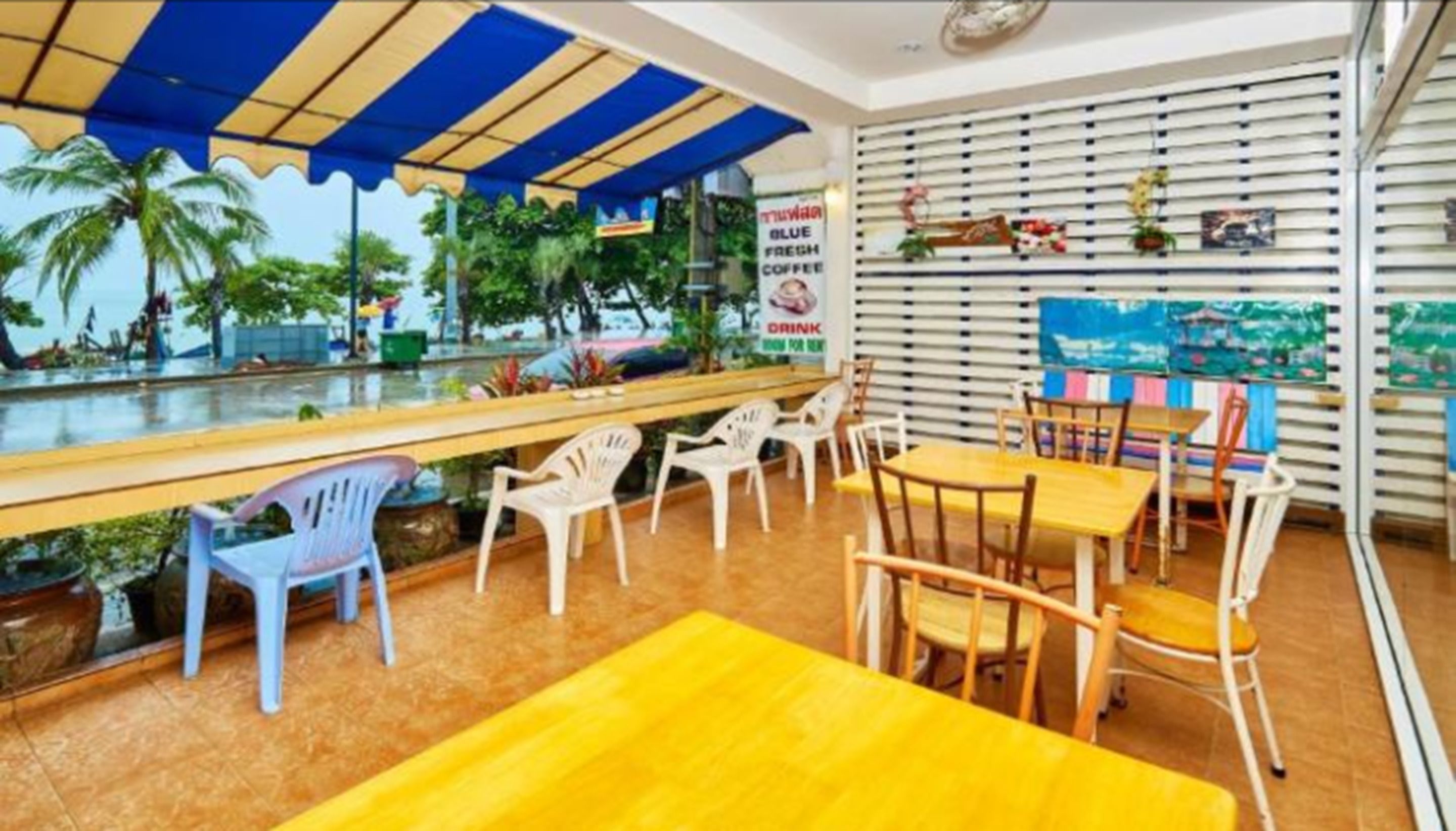 A café with outdoor seating, a blue and yellow striped awning, and a view of palm trees.
