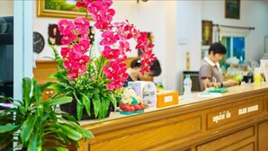 A reception desk with a wooden finish, a potted plant with pink flowers, and a clock on the wall. - Blue House Beach (Pattaya)