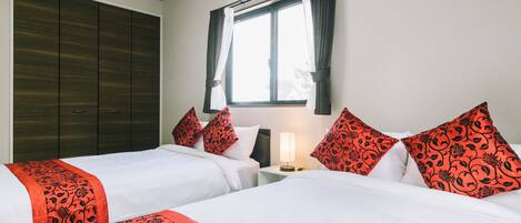 A hotel room with two beds, a wardrobe, a window with curtains, and a bedside table with a lamp.