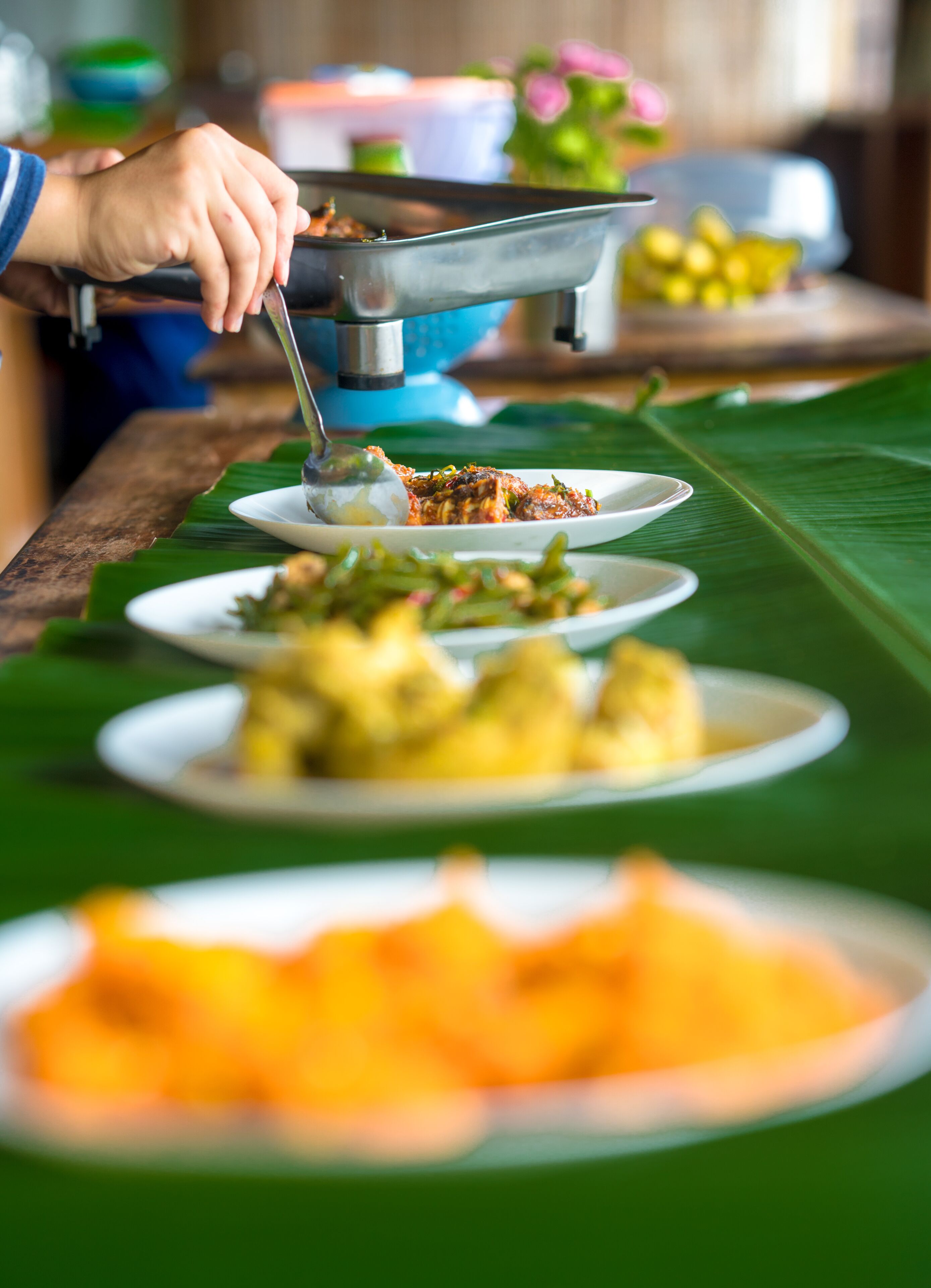 A buffet table with various dishes, including a plate with a yellowish food item, a plate with a green leafy dish, and a plate with a white food item.