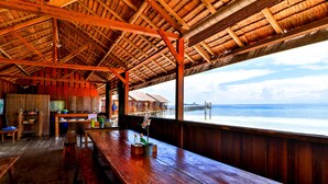 A wooden deck with a view of the ocean, wooden furniture, and a thatched roof structure. - HamuEco Dive Resort Raja Ampat (Waisai)
