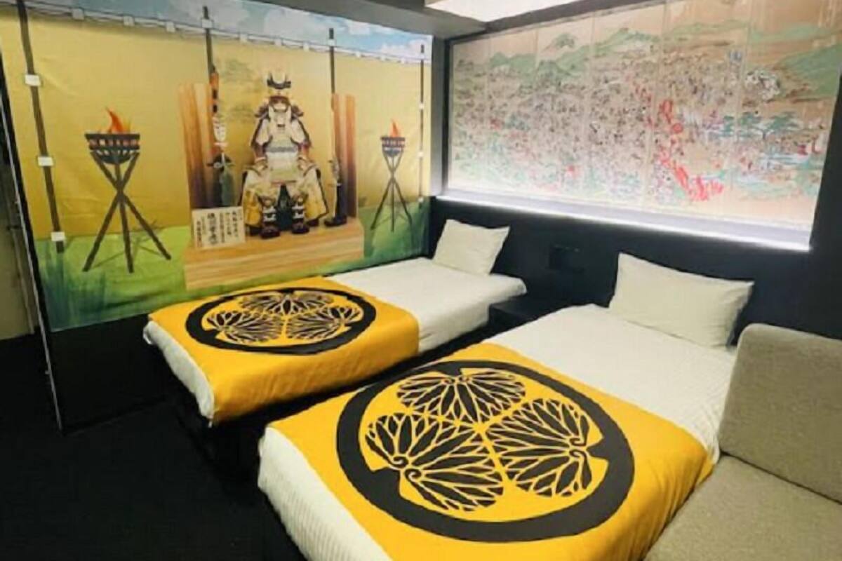A hotel room with two beds, each with a yellow and black patterned bedspread. There is a large map on the wall and a painting depicting a historical scene.