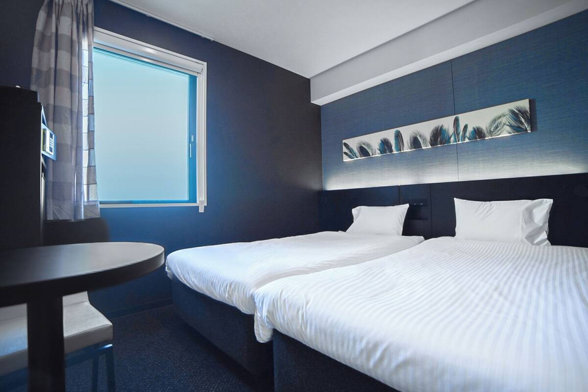 A hotel room with two beds, a small table, a window, and a wall-mounted artwork.