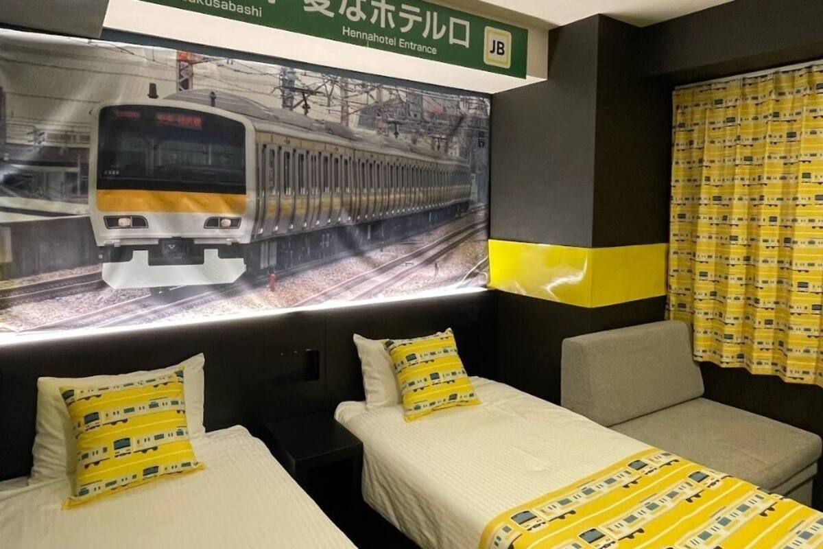 A hotel room with two beds, each with a pillow featuring a train pattern. A large window displays a train passing by. The room has a green sign above the beds with Japanese text.