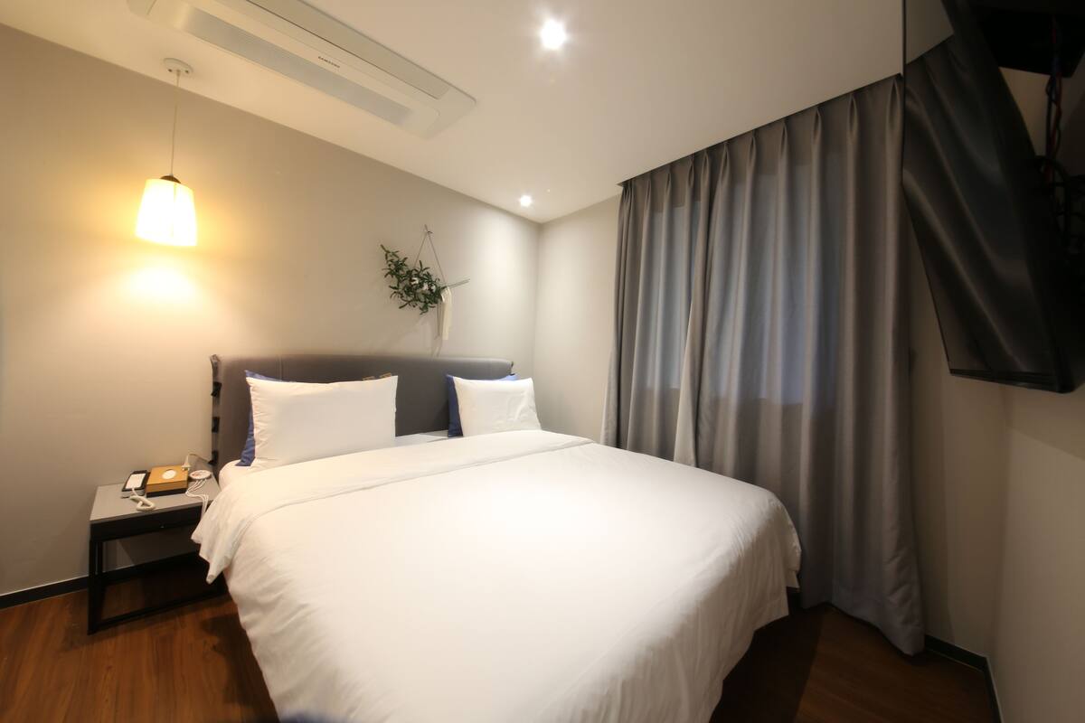 A neatly made bed with white linens, a bedside table with a phone and charger, a wall-mounted plant, and a large window with curtains.