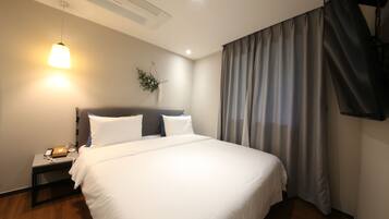 Una cama bien hecha con ropa de cama blanca, una mesita de noche con teléfono y cargador, una planta en la pared y un ventanal con cortinas.