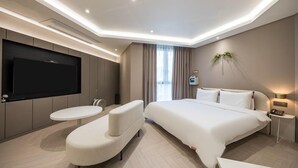 A modern hotel room with a large bed, a flat-screen TV mounted on the wall, a small round table, and a seating area with a cushioned bench. - Brown Dot Hotel Masan (Changwon)