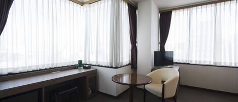 A hotel room with a desk, chair, TV, and large windows with curtains.