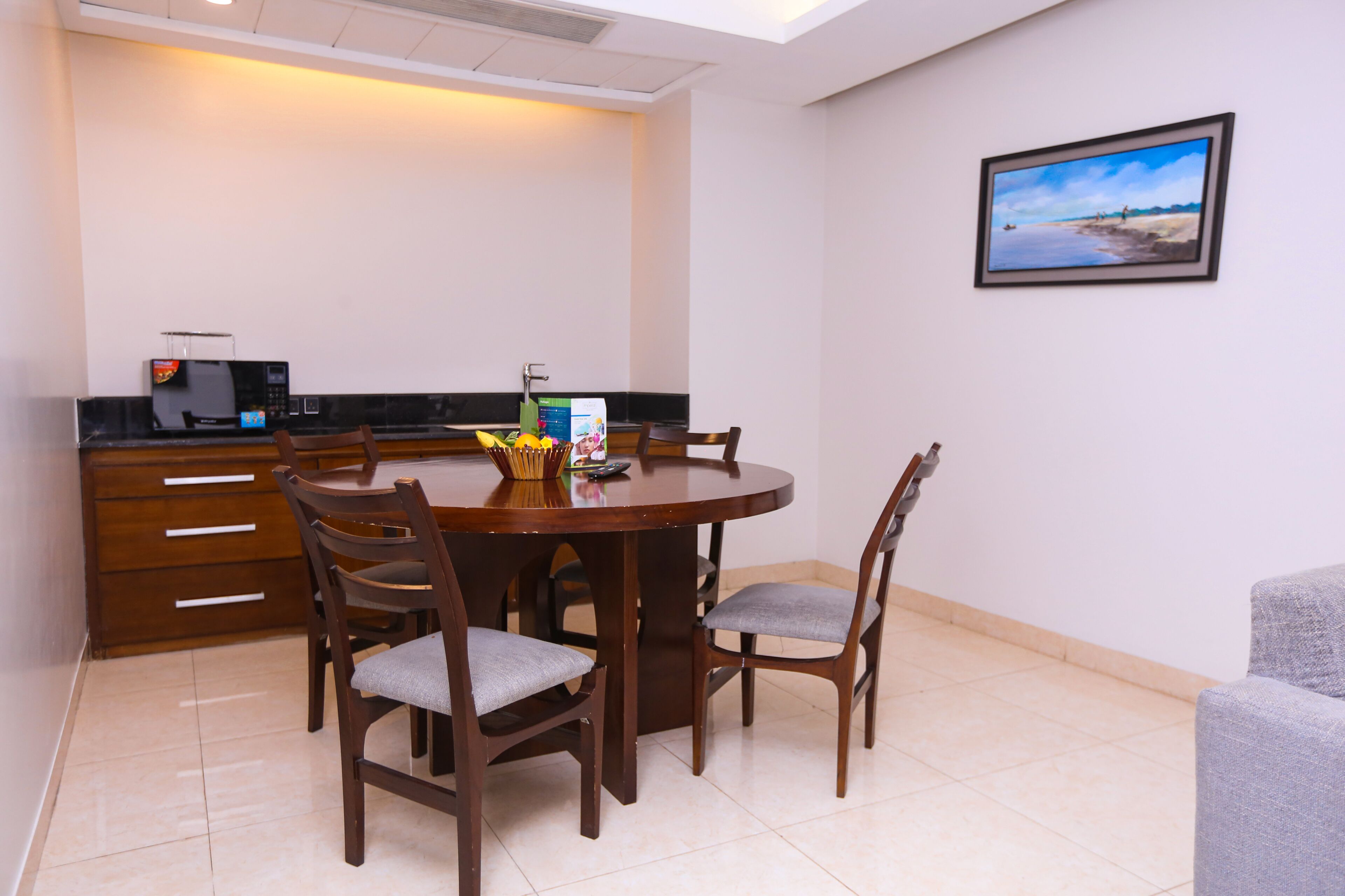 A dining area with a round wooden table, four chairs, a microwave, and a framed picture on the wall.