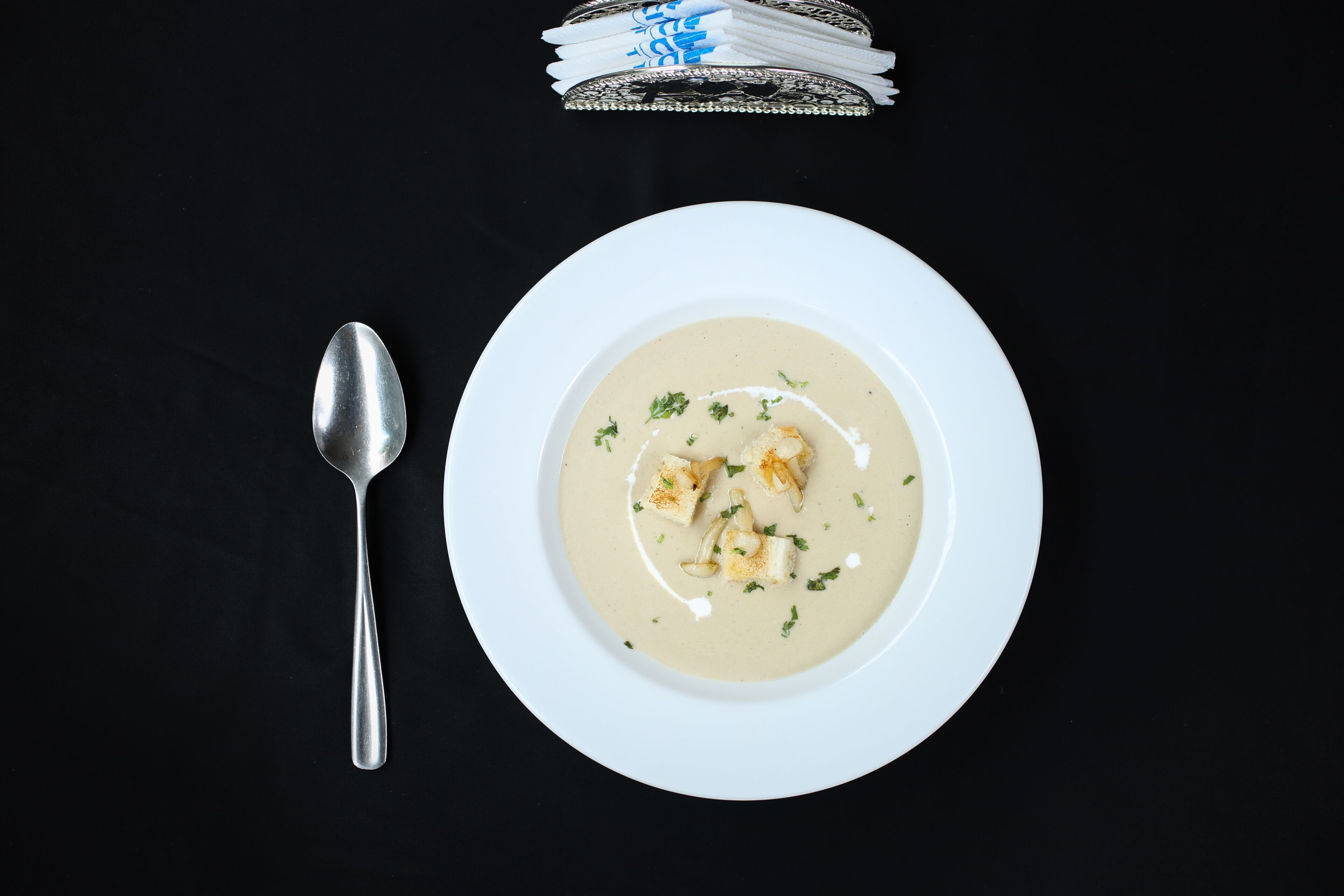 Um prato branco com sopa cremosa, decorado com ervas, uma colher e uma pilha de guardanapos dobrados sobre uma superfície preta.