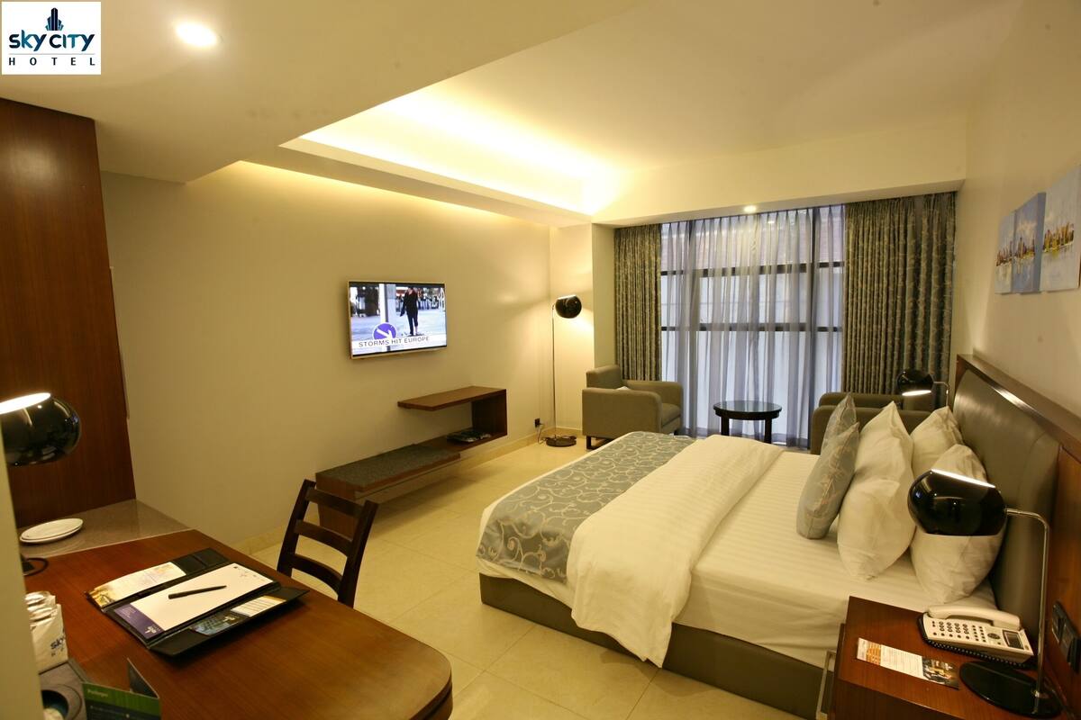 Habitación de hotel con cama, escritorio, silla, televisor y ventana con cortinas.