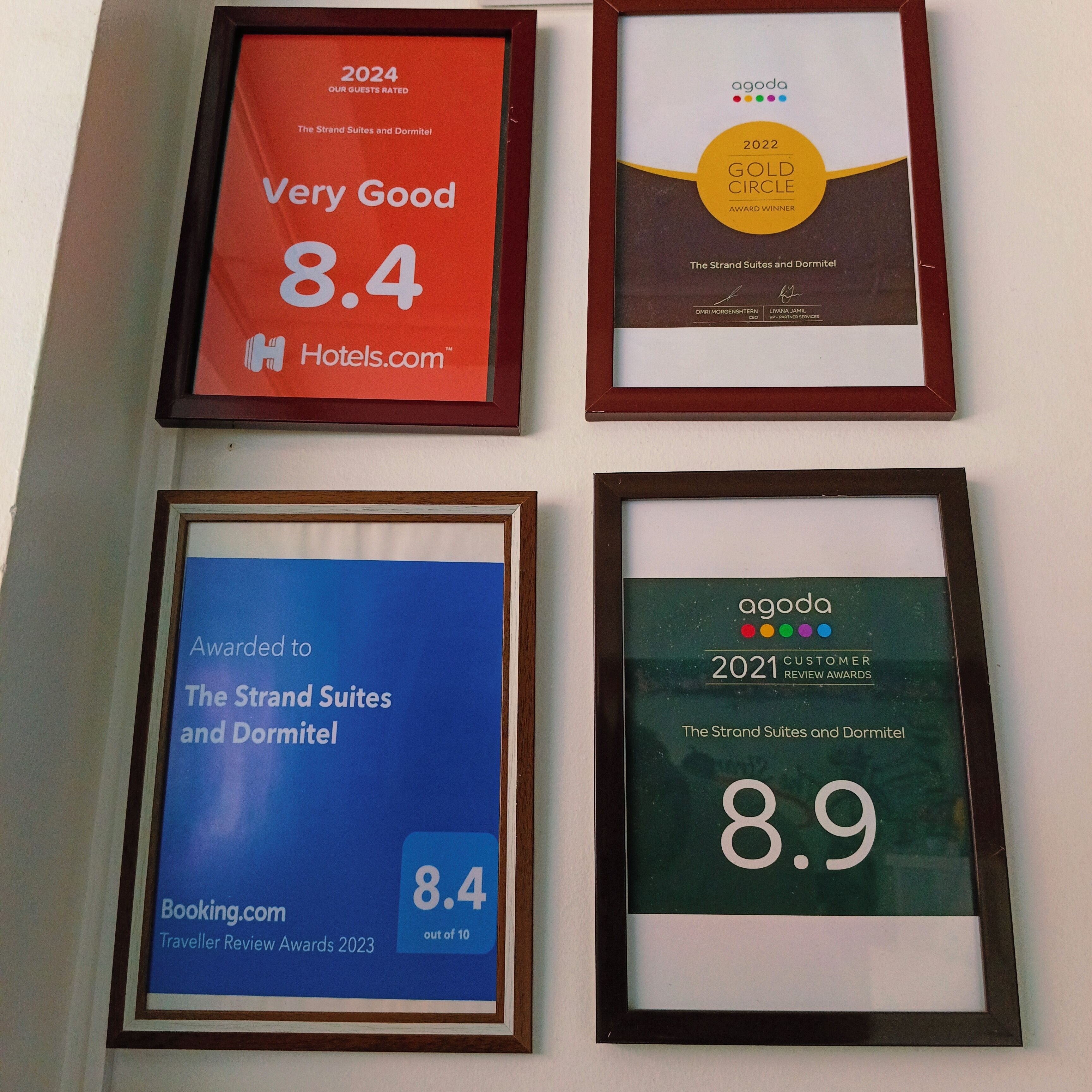 Quattro targhe incorniciate per The Strand Suites e Dormitel. Ogni cornice mostra un premio diverso da Hotels.com, Booking.com e Agoda, con valutazioni di 8.4 e 8.9.