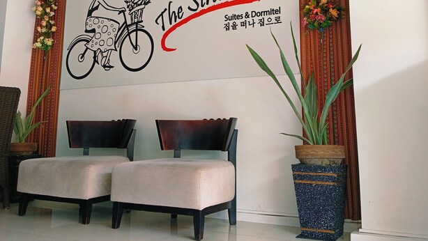 A reception area with a wall-mounted sign reading "The Strand Suites & Dormitel." There are two upholstered chairs in front of the sign, a potted plant to the right, and a decorative garland above the sign. - The Strand Suites and Dormitel (Davao)